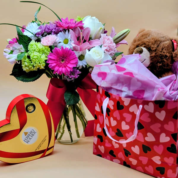 Mixed bouquet in a glass vase beside a teddy bear gift bag and heart-shaped chocolate box