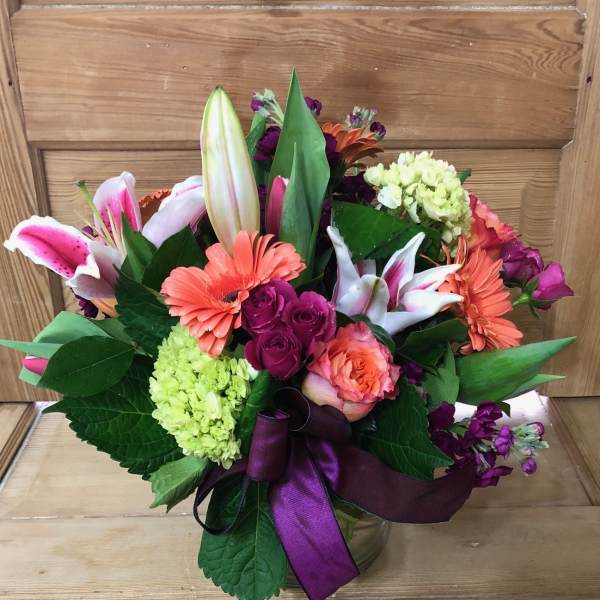 Mixed bouquet of lilies, roses, and gerbera daisies in a glass vase with a purple ribbon