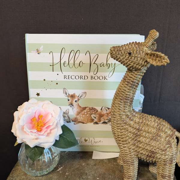 Single pink-and-white flower in a small glass vase beside a baby record book and woven deer figurine.