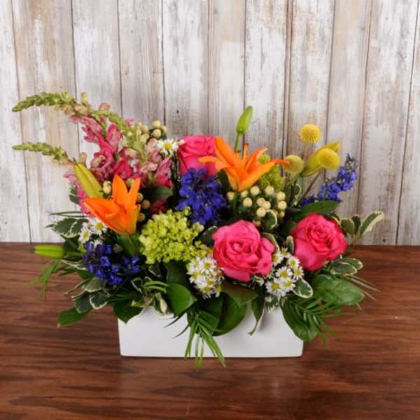 Low rectangular arrangement of bright pink roses, orange lilies, blue flowers, and mixed blooms in a white container