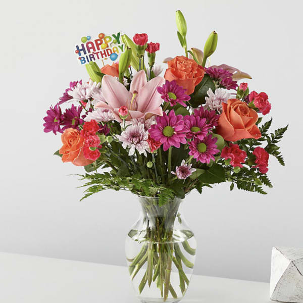Mixed bouquet of pink lilies, roses, and daisies in a glass vase with a birthday pick