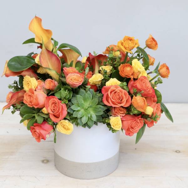 Low arrangement of orange and yellow roses, calla lilies, and a green succulent in a white cylinder vase