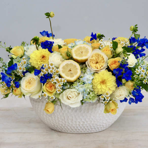Low yellow, white, and blue flower arrangement with lemon slices in a white ceramic bowl