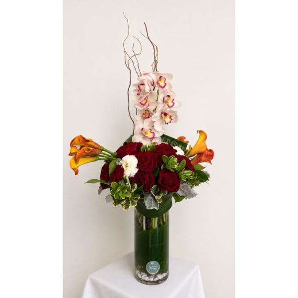 Tall floral arrangement with red roses, white orchids, and orange calla lilies in a glass vase.