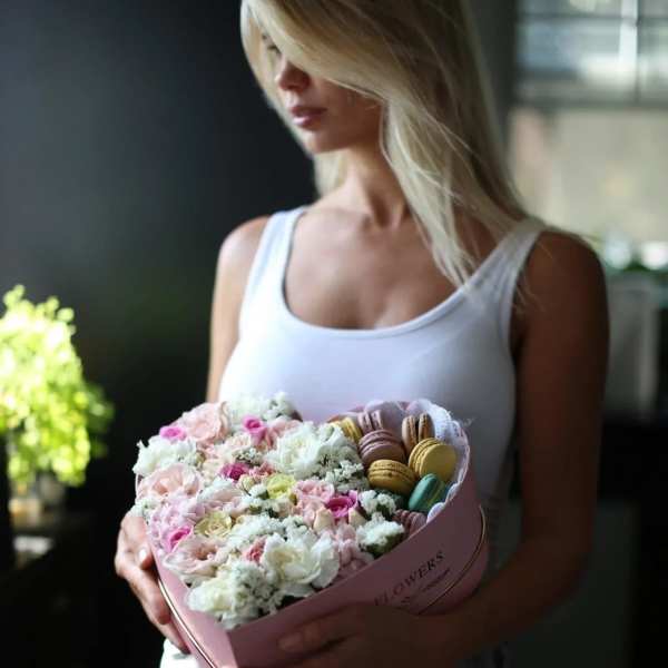 Woman holding a pink heart-shaped box of pastel flowers and macarons
