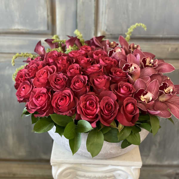 A low arrangement of red roses and burgundy orchids in a white bowl.