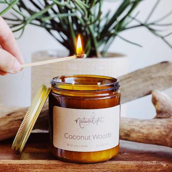 Amber glass jar candle labeled Coconut Woods being lit with a wooden match.