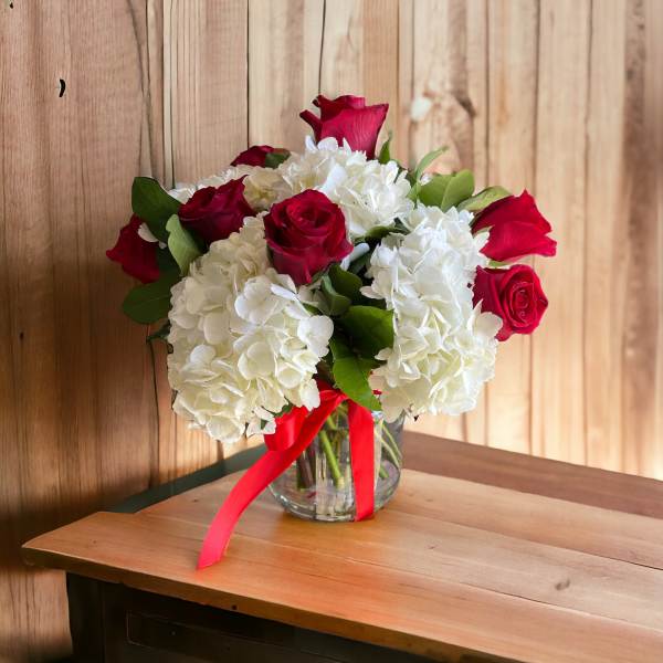 Arrangement of red roses and white hydrangeas in a clear glass vase with a red ribbon.