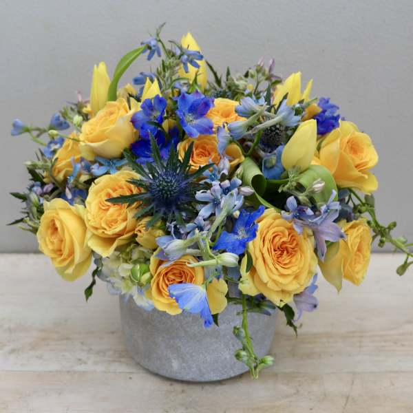 Low arrangement of yellow roses and tulips with blue flowers in a gray pot