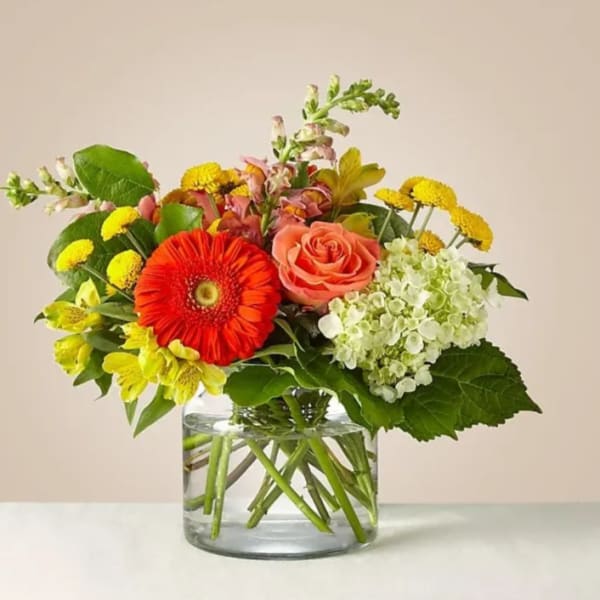 Mixed bouquet with a coral rose, red gerbera daisy, and yellow flowers in a glass vase