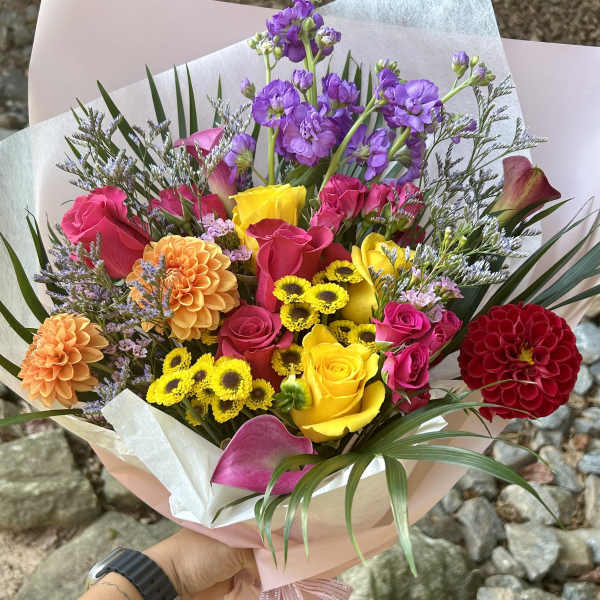 Handheld bouquet of multicolored roses, dahlias, and purple blooms wrapped in pink paper