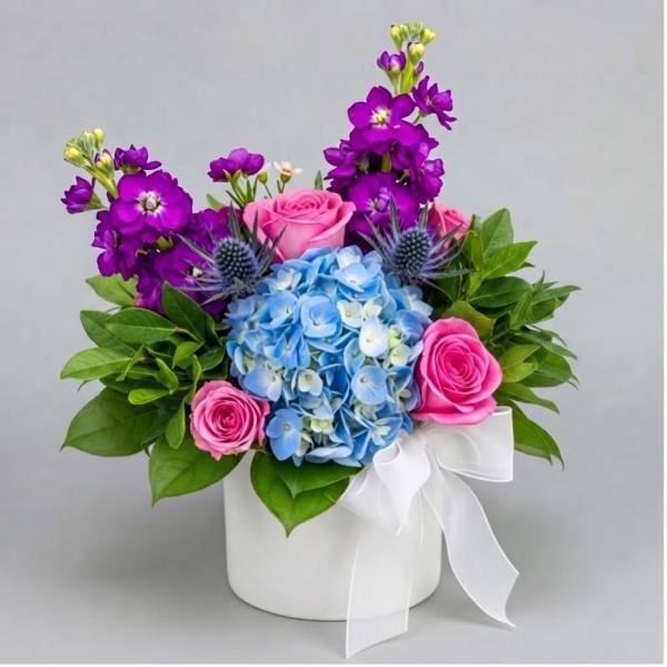 Bouquet of blue hydrangea, pink roses, and purple flowers in a white box with ribbon
