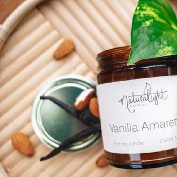 Brown glass Vanilla Amaretto candle jar with white label on a tray beside almonds, vanilla beans, and a green vine.