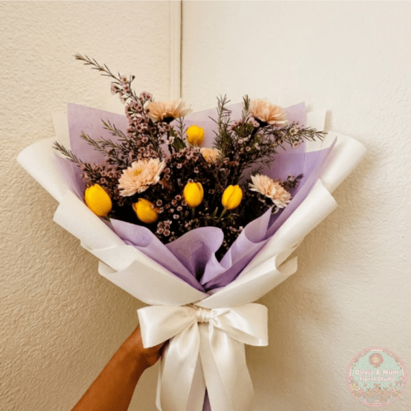 Handheld bouquet of yellow tulips and pale flowers wrapped in lavender and white paper