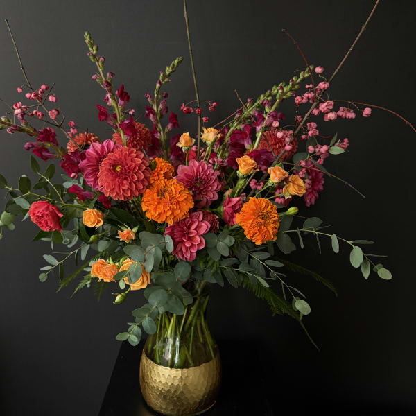 Tall arrangement of orange marigolds, pink dahlias, and spray roses in a glass vase with a gold base.