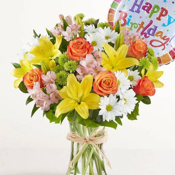 Bouquet of yellow lilies, orange roses, and white daisies in a glass vase with a birthday balloon