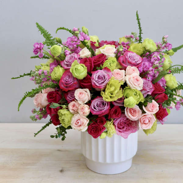 Lush arrangement of pink, red, lavender roses and green blooms in a white vase