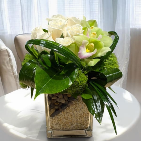 White roses and pale green orchids in a square glass vase with decorative stones.