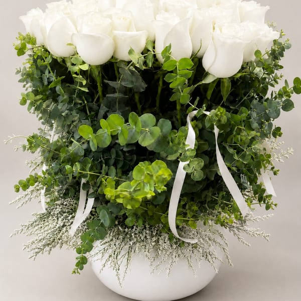 White roses arranged in a round white vase with lush greenery and ribbon accents