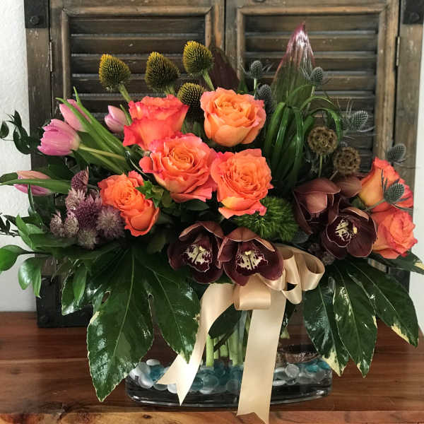 Mixed bouquet of orange roses, pink tulips, and dark orchids in a glass vase
