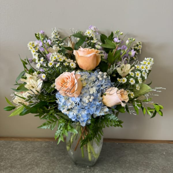 Mixed bouquet of peach roses, blue hydrangea, and small white daisies in a glass vase