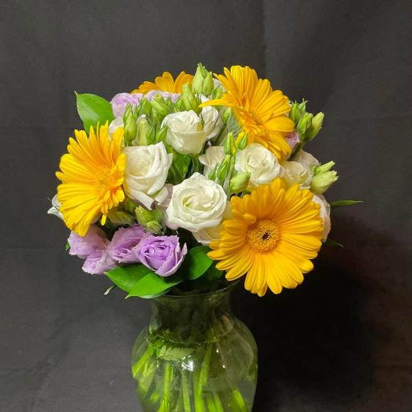 Bouquet of yellow gerbera daisies, white roses, and lavender blooms in a glass vase