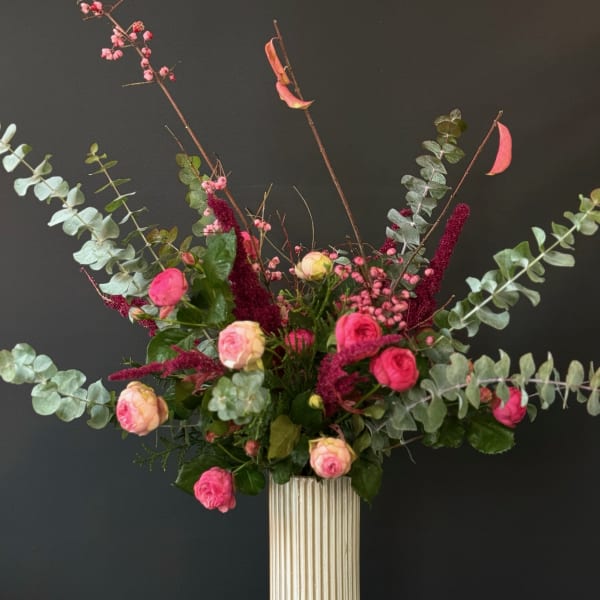 Tall arrangement of pink roses, berries, and burgundy accents in a white ribbed vase with sweeping eucalyptus.