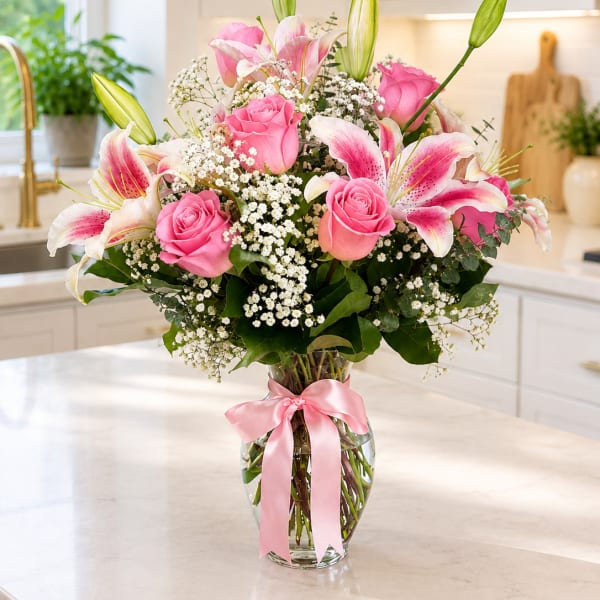 Pink roses and lilies arranged in a clear glass vase with a pink ribbon.
