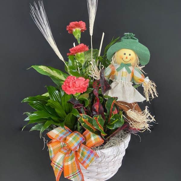 Basket arrangement with pink carnations, tropical foliage, and a scarecrow doll