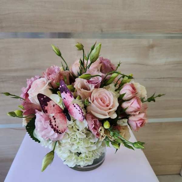 Low arrangement of blush pink roses and lisianthus with white hydrangeas in a glass vase and a pink butterfly accent