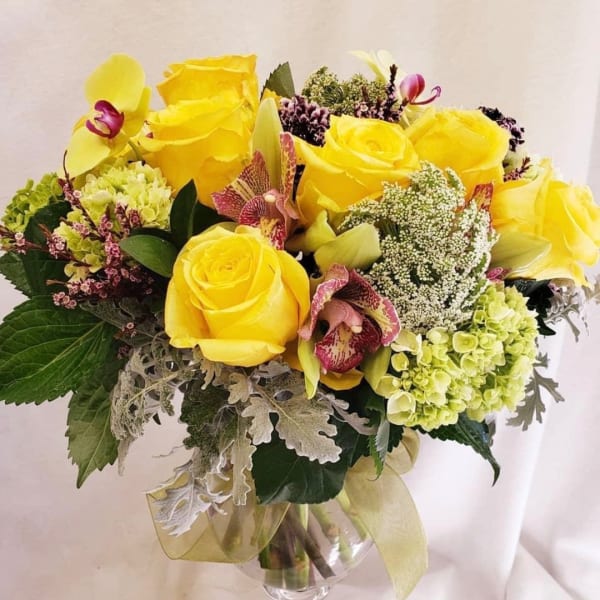 Yellow roses and orchids arranged in a clear glass vase with ribbon.