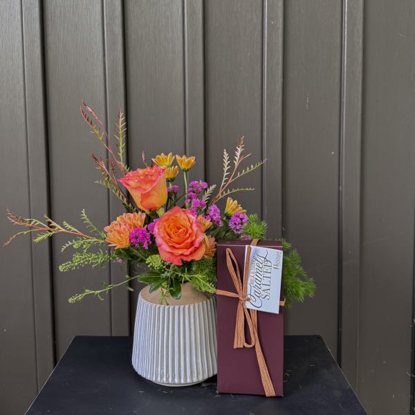 Orange roses and small purple flowers in a ribbed vase beside a boxed chocolate bar