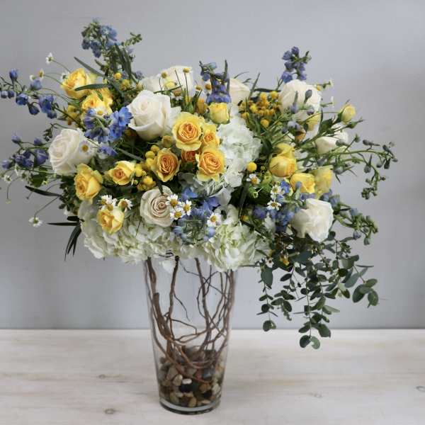 Tall arrangement of yellow and white roses, blue flowers, and hydrangeas in a clear glass vase.