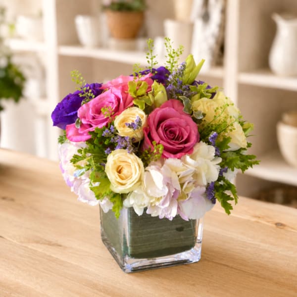 Mixed bouquet of pink, cream, and purple flowers in a square glass vase