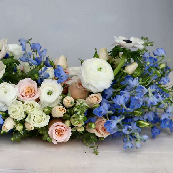 Low centerpiece of white ranunculus, blue delphinium, and blush roses in a long horizontal arrangement