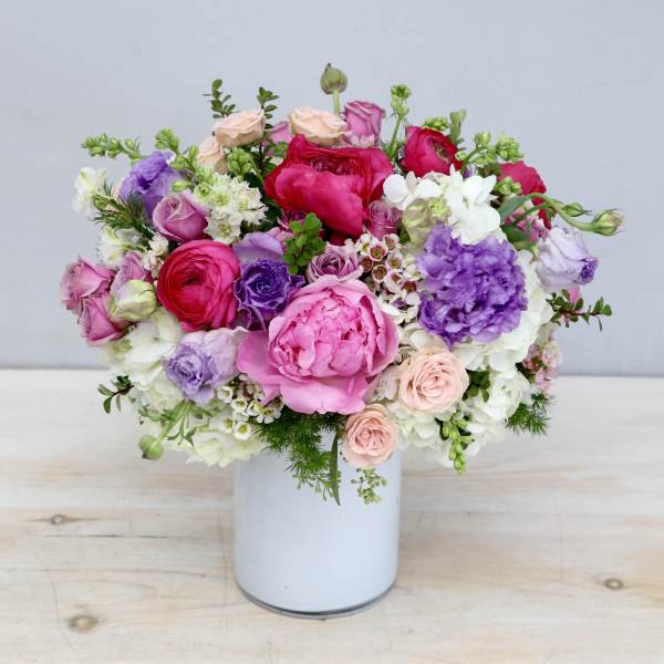 Round arrangement of pink peonies, roses, and purple blooms in a white vase