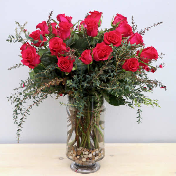 Tall arrangement of bright red roses with abundant greenery in a clear glass vase with pebbles.