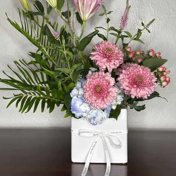 Pink lilies and chrysanthemums arranged in a white gift box with blue hydrangea.