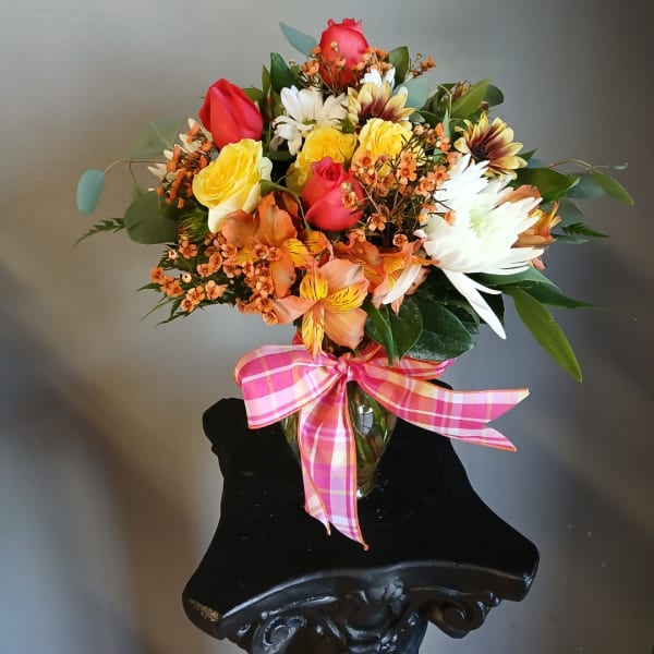 Mixed bouquet of roses, daisies, and orange flowers in a glass vase with a pink plaid ribbon
