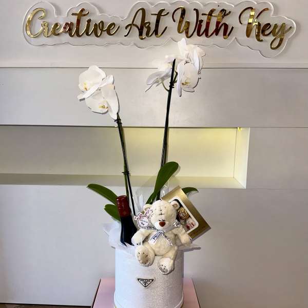 White orchids in a glittery box with a teddy bear and wine bottle