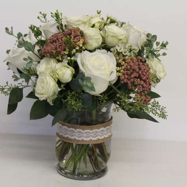 White roses and pink filler flowers in a glass vase with lace ribbon