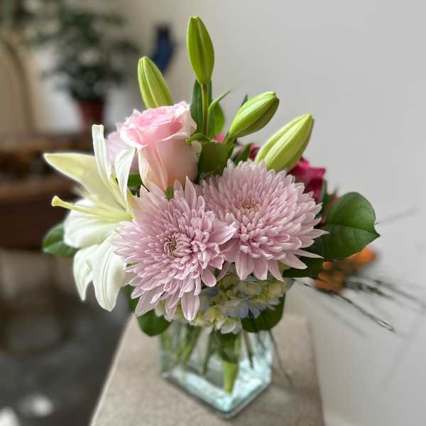 Pastel arrangement of pink chrysanthemums, a pink rose, and white lilies in a clear glass cube vase.