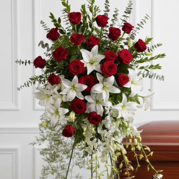 Tall standing spray of red roses and white lilies with trailing white orchids on a metal easel beside a wooden casket