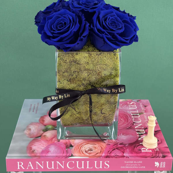 Three blue roses in a clear square vase with moss and a black ribbon.