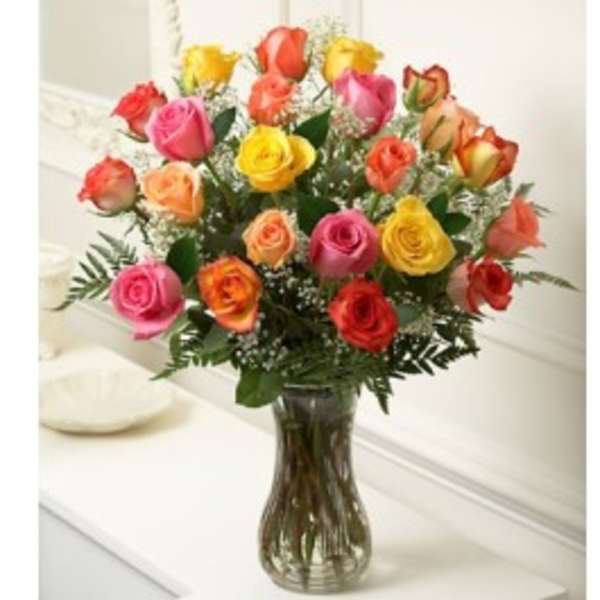 Tall glass vase filled with assorted pink, yellow, orange, and red roses with white filler flowers.