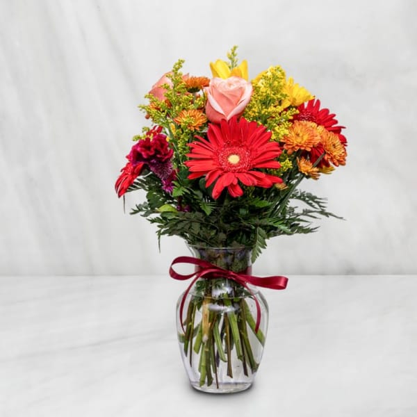 Mixed bouquet of red, pink, and yellow flowers in a glass vase with a ribbon