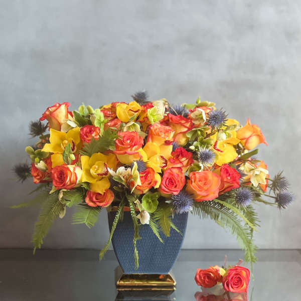 Low arrangement of yellow orchids and orange roses with blue accents in a blue vase with gold base