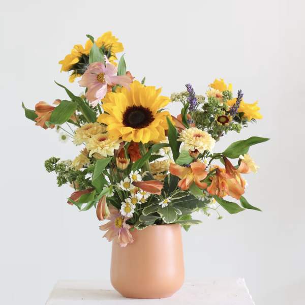 Mixed bouquet with sunflowers, orange lilies, and daisies in a peach vase
