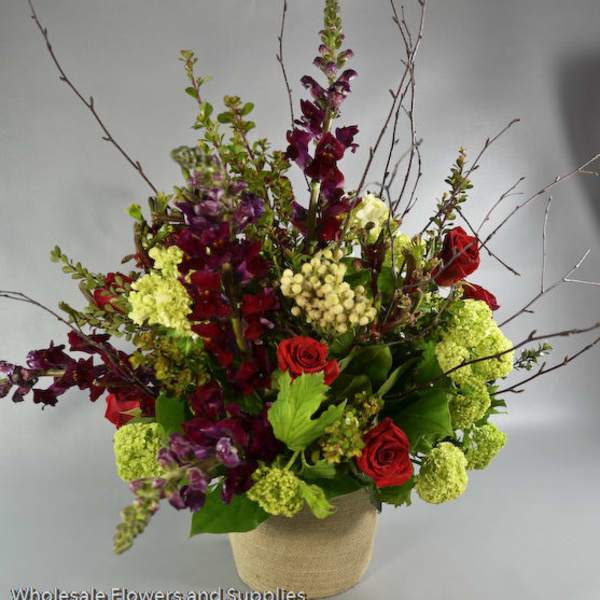 Tall floral arrangement with red roses, purple blooms, and green hydrangeas in a burlap-wrapped vase
