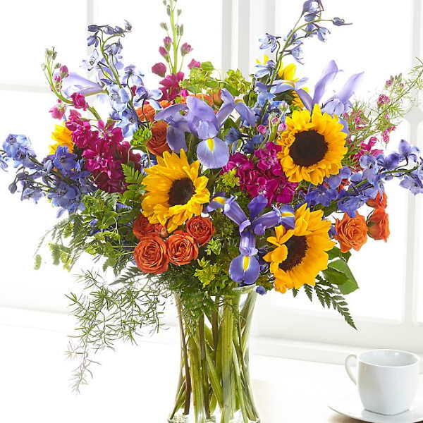 Colorful mixed bouquet in a clear glass vase with sunflowers, roses, and blue flowers